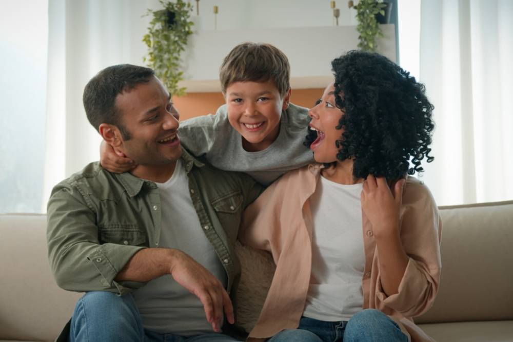 Affectionate parents adult African American mother and father embrace cuddle on couch funny little boy adopted son