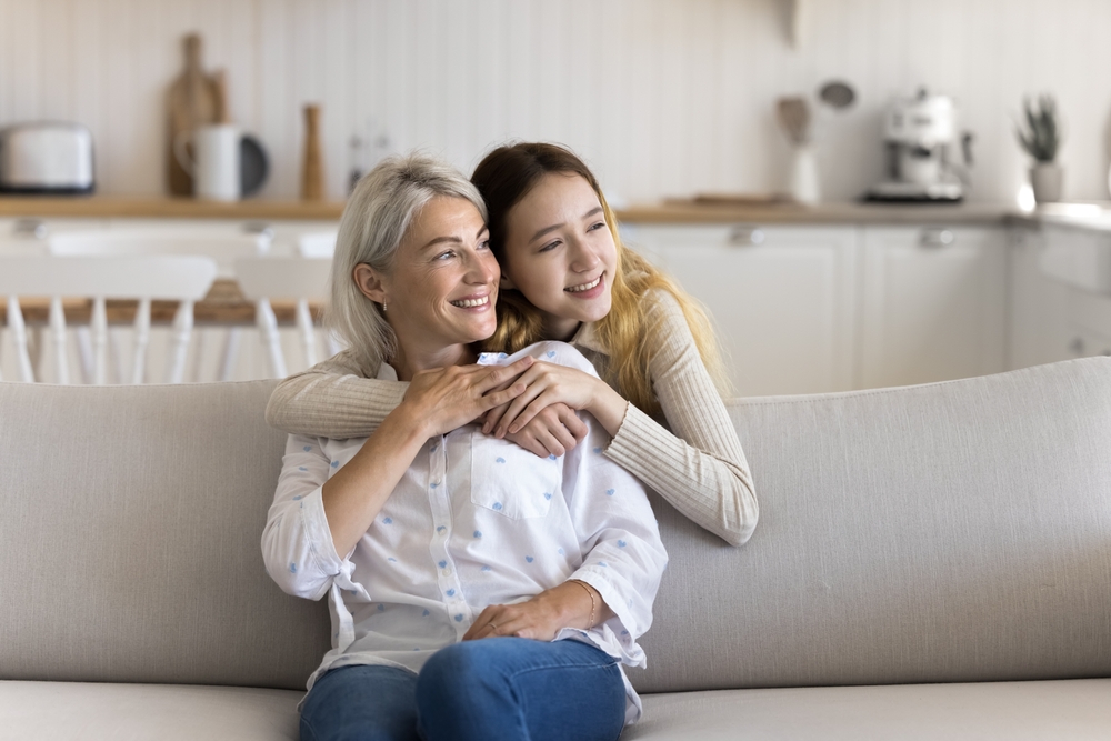 Positive dreamy teenage kid hugging mature grandma from behind at home, looking away, smiling, thinking